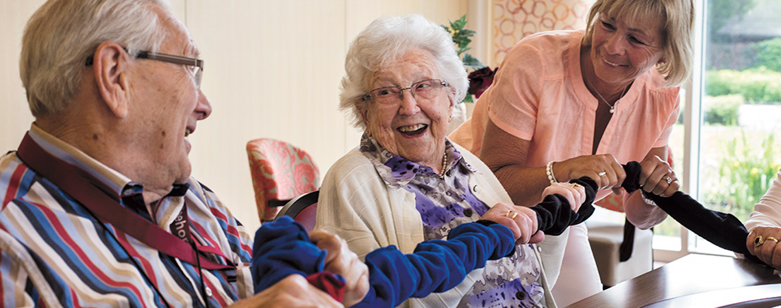 Auf dem Foto sehen Sie zwei ältere Menschen mit Demenz zusammen mit einem Aktivitätsbetreuer, die mit dem Kooperationsband von SpielePlus, einem Laden speziell für Spiele für Senioren, spielen.